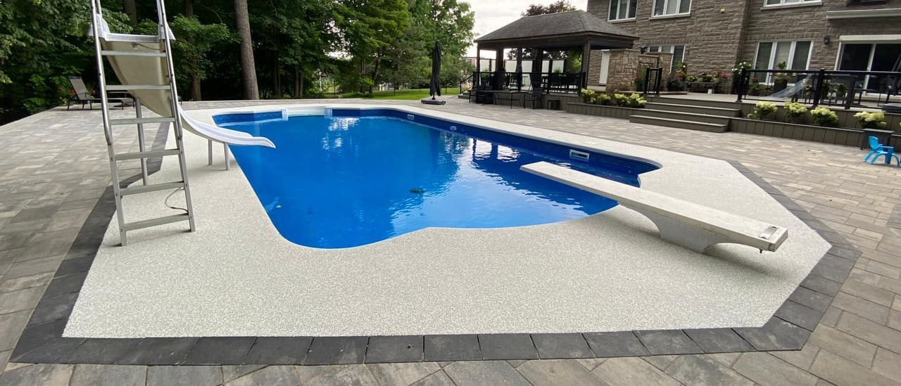 A large pearl-white Gator Grip pool deck with a charcoal border, surrounding an in-ground pool at a stone country home